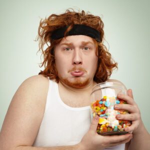 Portrait of funny overweight young redhead male with sports band on his head having unhappy and sad look, holding glass jar of sweets, longing for unhealthy carbohydrates after physical exercises