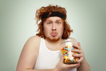 Portrait of funny overweight young redhead male with sports band on his head having unhappy and sad look, holding glass jar of sweets, longing for unhealthy carbohydrates after physical exercises