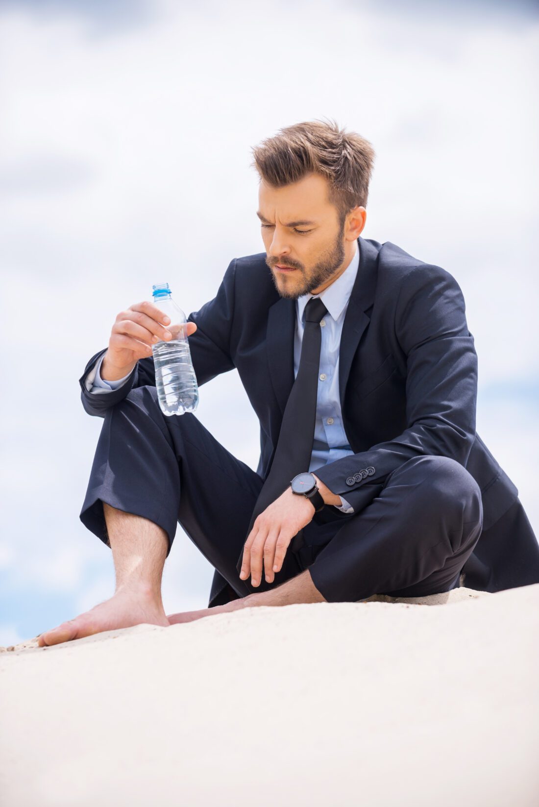Water is life. Depressed young businessman holding bottle with water and looking at it while sitting on sand