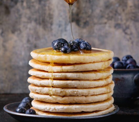 Pancakes with fresh blueberries in blue ceramic plate, and flowing honey from wooden honey dipper, served on black iron surface with tin textured background.