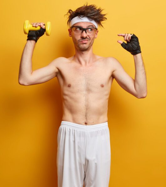 Photo of unimpressed European man shows small gesture, tells how much he worked on muscles, lifts dummbell, wants to have perfect biceps, wears sport gloves, poses against yellow background free space