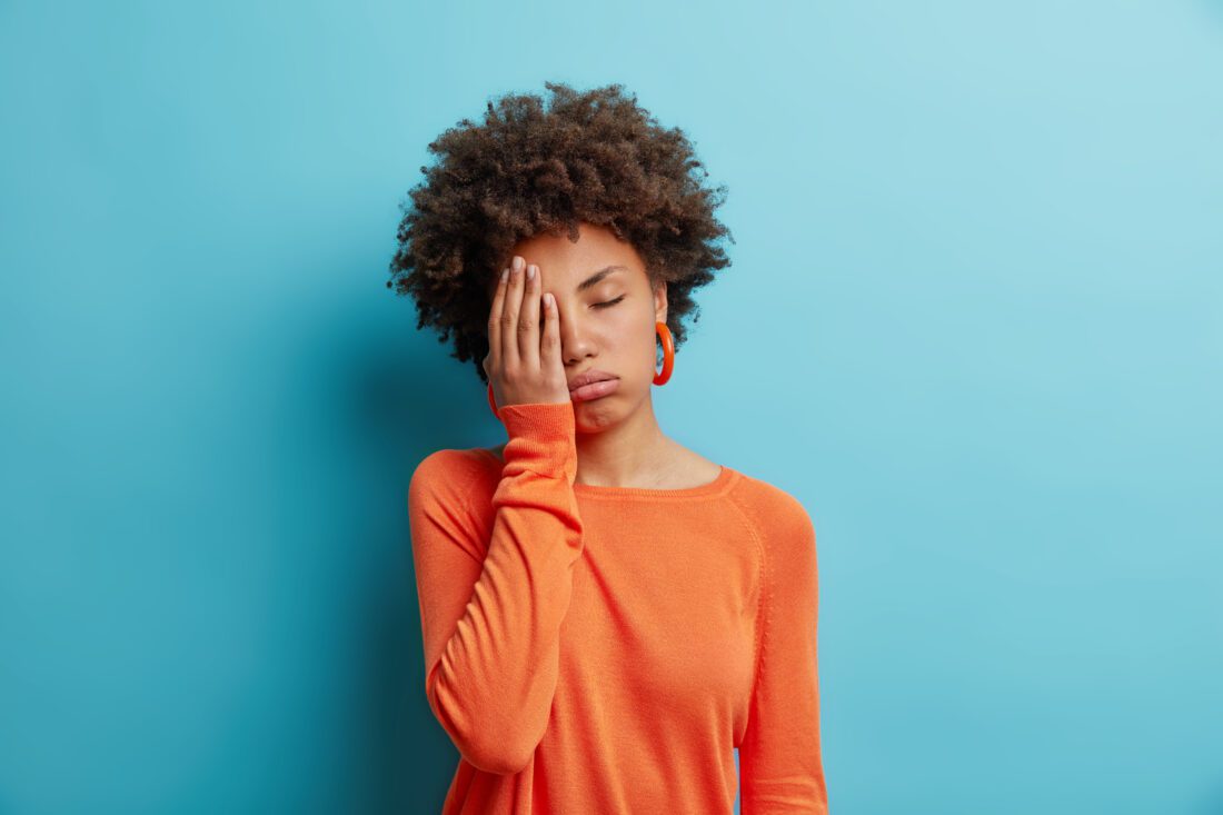 Exhausted unhappy woman makes face palm and sighs from tiredness has sleepy expression fed up of working without rest wears orange jumper in one color with earrings. Upset depressed female model this can be an obstacle to exercise